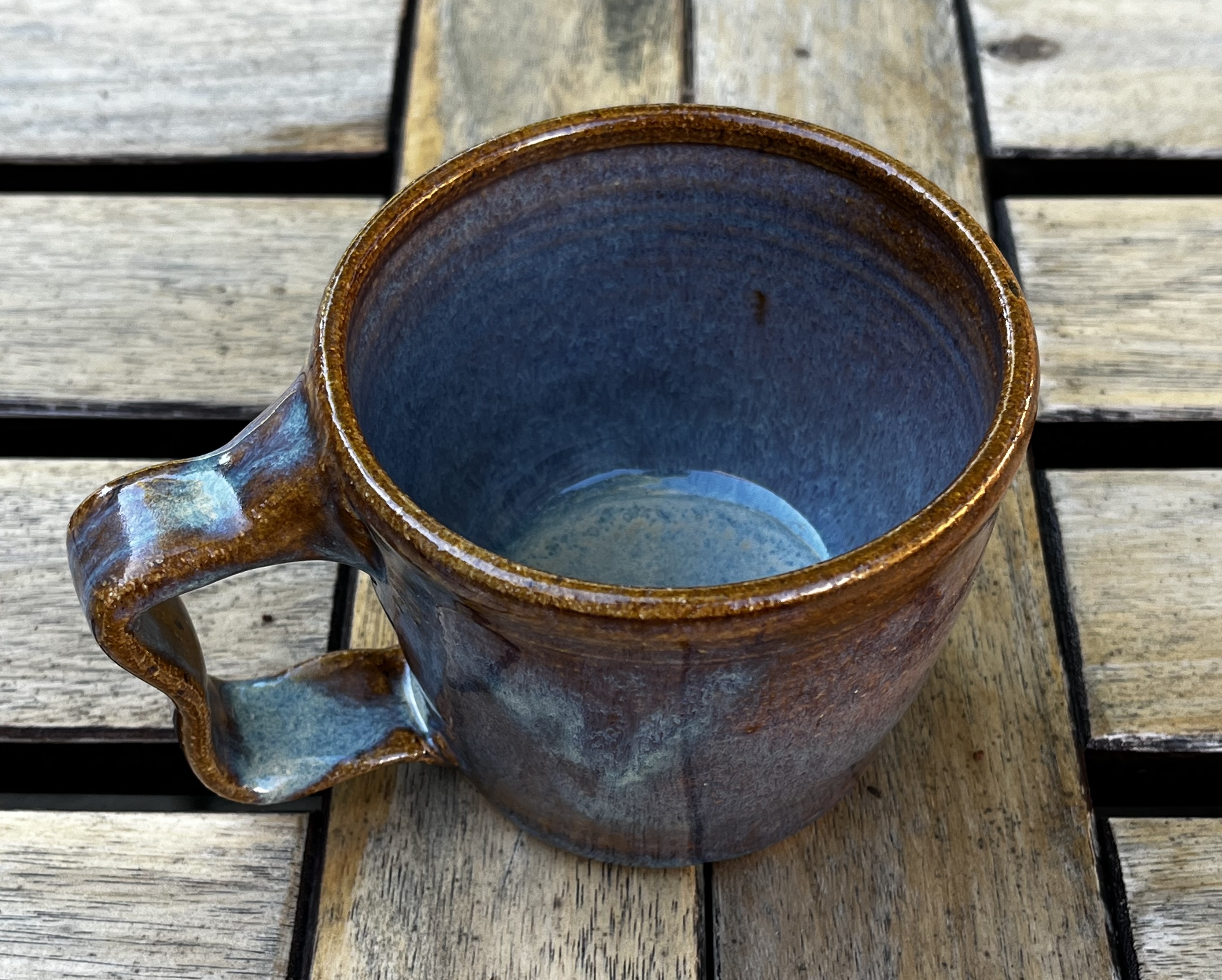 A mug with wiggly handle and brownish glaze with pops of blue all along the inside and a bit on the handle and sides.