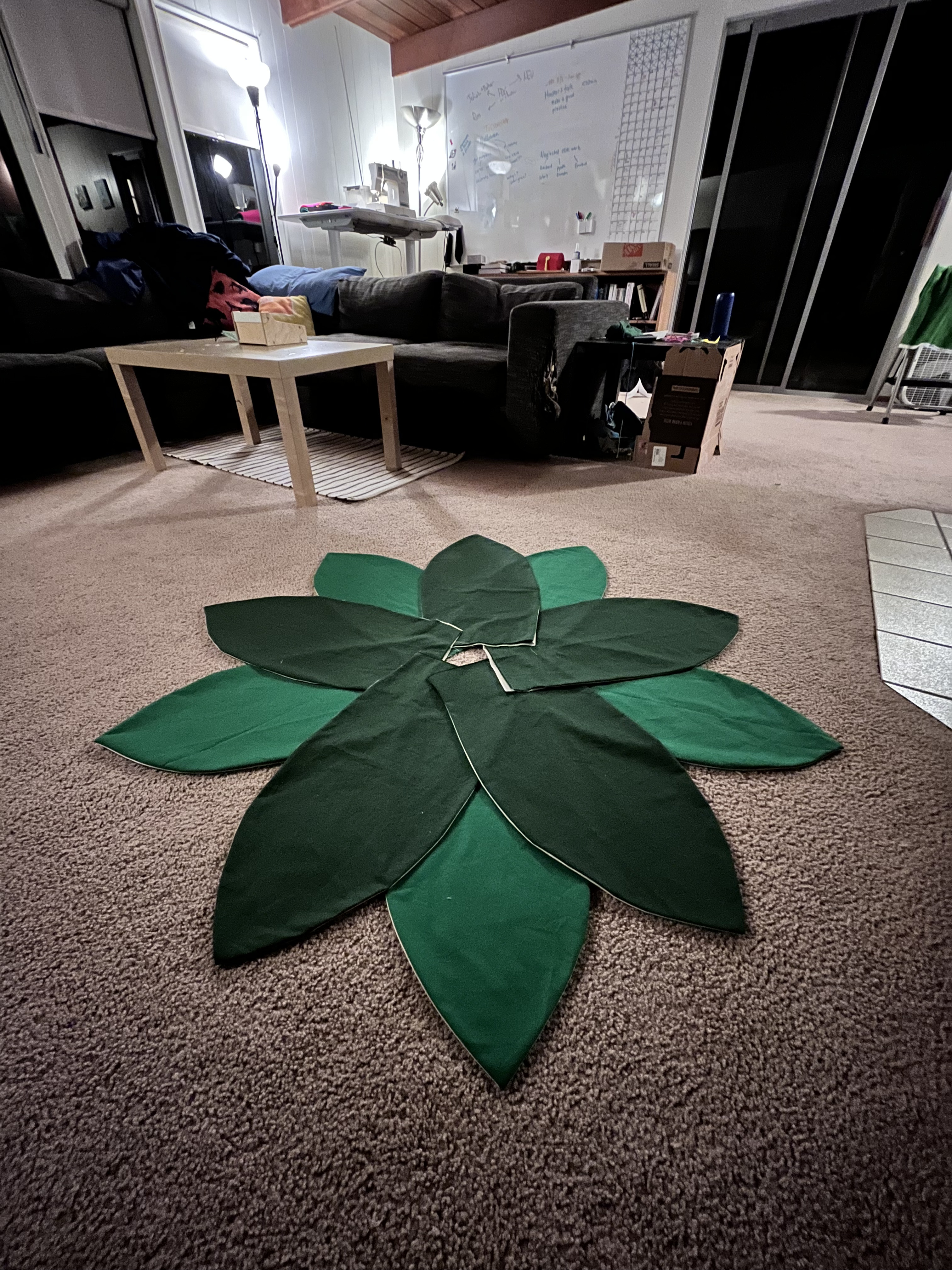 Dark and lighter green leaf fabric shapes laid out in a circle on a carpeted floor in a dim living room